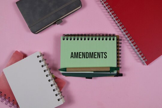 a spiral notebook with the word &ldquo;Amendments&rdquo; surrounded by colorful notebooks and a pen on an pink background, representing legal changes, document revision, policy updates, and business planning.
