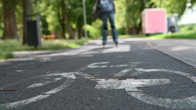 Blonde teen girl rollerblades paved park path. Wears hoodie, jeans, backpack. Green trees visible.