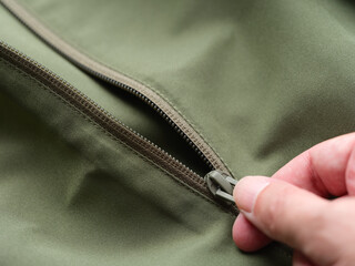 Close-up of a person's hand pulling the zipper slider on a green fabric jacket. © Stepan Popov