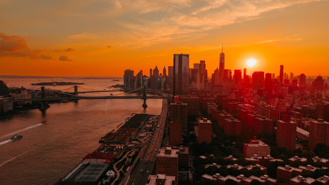Manhattan Bridge and city skyscrapers during vibrant orange sunset in New York. Setting sun glows bright orange behind urban skyscrapers and bridges along East River.