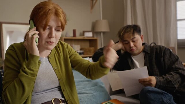 Caucasian young adult woman talking on smartphone and gesturing stressed over bills while Asian young adult man sitting behind. Ideal for banking, debt relief, or financial planning ads