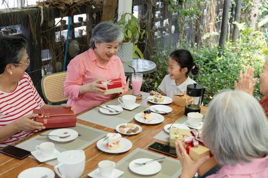 Retired friends and a granddaughter enjoy gift giving at an outdoor cafe, sharing tea, cakes and friendly conversation while smiling during a relaxed reunion with warm companionship, aging society.
