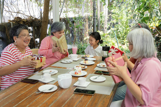 Longtime friends and a child exchange wrapped gifts at an outdoor cafe, reaching across the table as tea and cakes set the scene for a joyful reunion with delight and warm connection, aging society.