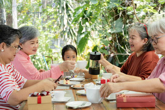Senior women and a young girl hold gift boxes at a garden cafe, enjoying a playful reunion with desserts, tea and bright laughter as they share happy surprises and lively conversation, aging society.