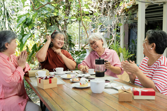 Old friends gather around a garden cafe table, laughing and clapping during a cheerful reunion with tea, cake and conversation that reflects friendship and joyful connection for all, aging society.