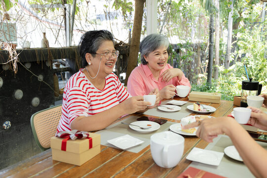Retired friends enjoy tea and desserts at a garden cafe, laughing around the table during a lighthearted reunion that captures friendship, relaxation, easy conversation and shared joy, aging society.