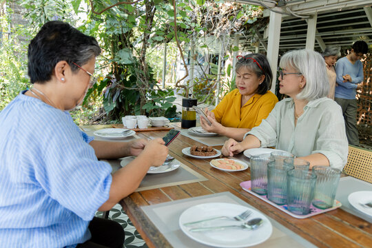 Retired friends sit together at a garden cafe, each checking a mobile phone while chatting, reflecting digital lifestyle, companionship and everyday social connection in later life, aging society.