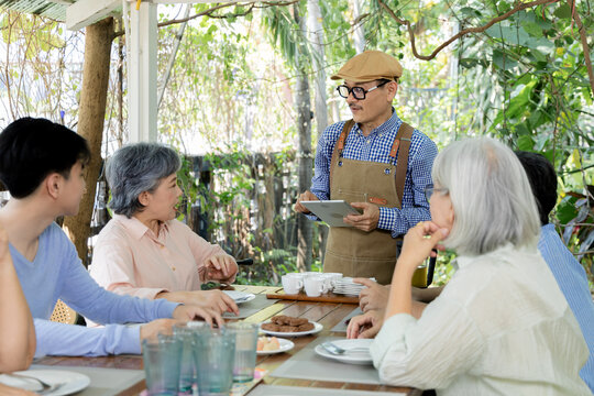 A cafe waiter stands with a tablet beside senior friends and a young man at an outdoor table, creating a candid moment of ordering, conversation and warm intergenerational connection, aging society.