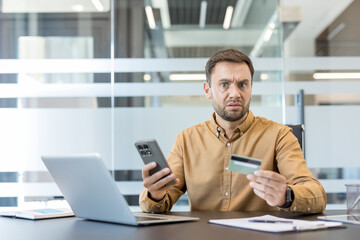 Stressed man looking at the camera with a surprised expression, holding a smartphone and credit...