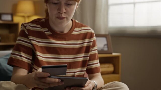Caucasian young adult woman holding credit card and entering details on tablet, camera tilting down to reveal past due bills. Ideal for online banking and debt relief services