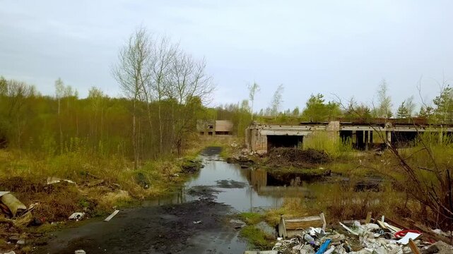 Abandoned and ruined brick building surrounded by mud, puddles, and piles of garbage under gloomy overcast sky, representing environmental pollution and decay