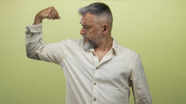 Middle aged man flexing bicep in studio against light green backdrop, rolled sleeve and visible forearm muscle; confidence strength.