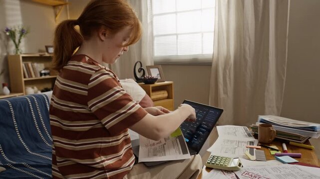 Young adult Caucasian woman sitting on sofa, reviewing utility bills and typing on laptop. Ideal for financial planning, debt relief, or online banking services commercials