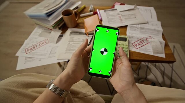 Unrecognizable woman holding smartphone with green screen in POV shot over table with past due bills. Ideal for showcasing mobile banking apps, debt relief services, or financial planning software