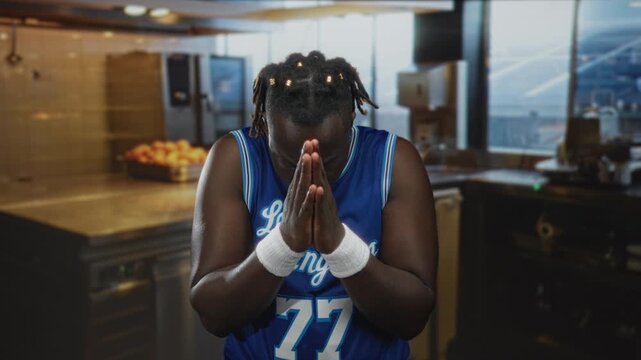 Man in a blue basketball jersey number 77 with white wristbands praying with hands pressed together and head bowed in a kitchen inside a building; solemn reflection.