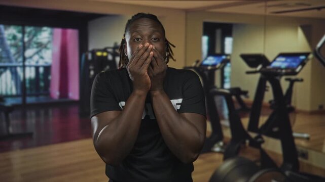 Man covering mouth with both hands while facing camera in gym with exercise bikes and mirrors visible; surprise silence.