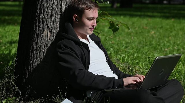 Young attractive caucasian sitting at the city park and working on his laptop computer. it startup programmer man working in the park using laptop sitting on the grass in sunny day