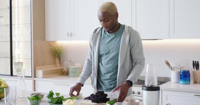 African American adult male reaching for herbs, arranging greens on board in kitchen for blender