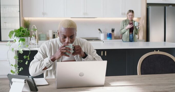 Diverse coworkers starting work session, man smiling, typing on silver laptop while holding mug