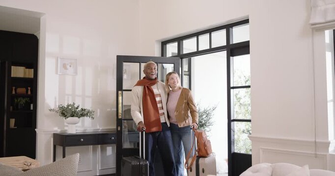 Diverse couple entering home after trip, man pulling black-framed glass door and carrying suitcases