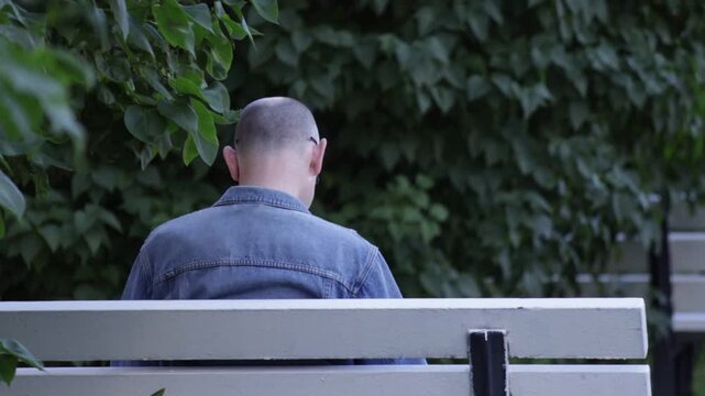 Man with shaved head in denim jacket sits on white bench