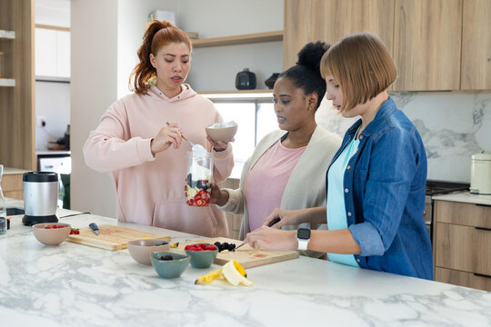 Diverse female friends preparing fruit yogurt blend on marble island with blender jar and knife