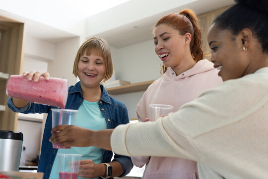 Three female friends pouring pink smoothie from pitcher into plastic cups at kitchen with blender