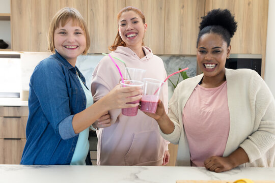 Diverse female friends standing at marble kitchen island smiling and clinking pink smoothie cups
