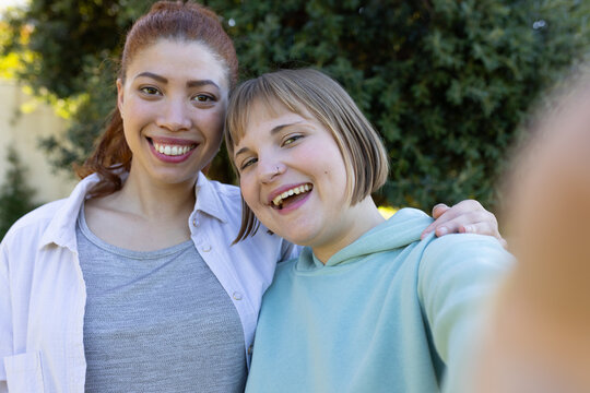 Two female friends smiling and leaning together in backyard, holding smartphone and wearing hoodie