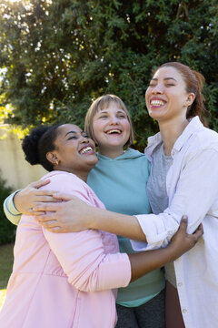 Diverse female friends hugging and laughing in sunlit backyard with light pink jacket, blue hoodie