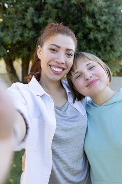 Diverse female friends taking selfie and leaning together outdoors near evergreen, wearing hoodie