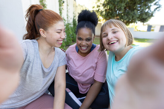 Diverse female friends leaning on bench in backyard in pastel activewear taking selfie with phone
