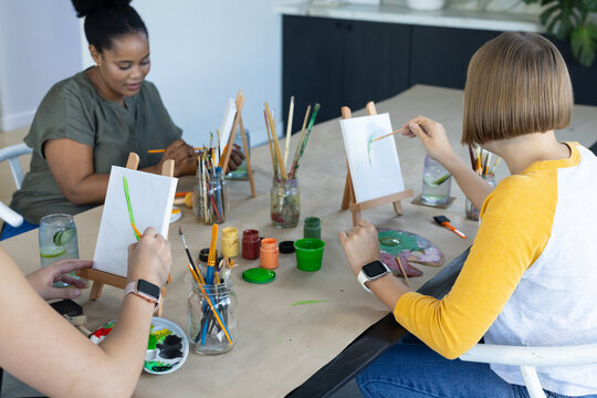 Diverse female friends painting on tabletop easels at studio table with brush jars, paint pots