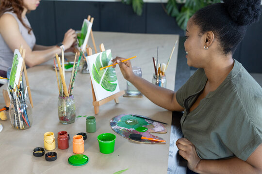 Two female painters sitting at craft table painting green leaf canvases with brushes, copy space