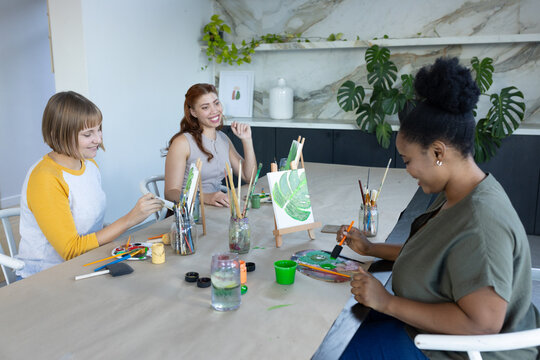 Diverse female friends in casual clothes painting and chatting at art studio with easel, copy space