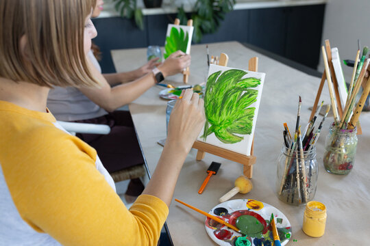 Diverse female friends painting green leaf motifs on small canvases at table with easel, brushes