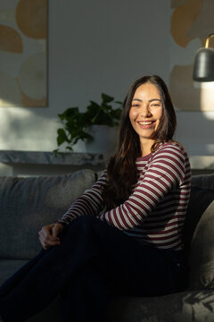Adult female sitting on gray sofa in living room, smiling, wearing maroon striped top, plant nearby
