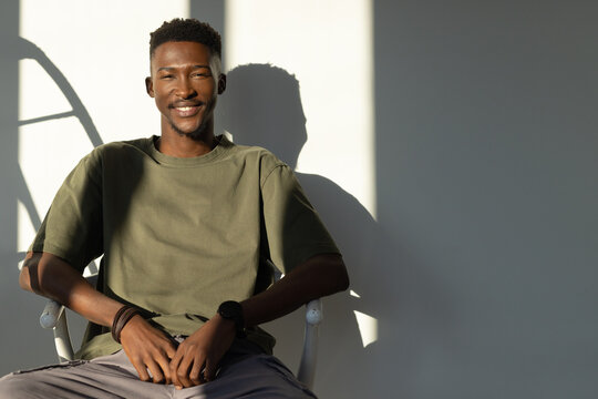 Male sitting smiling in olive T-shirt on white metal chair by light wall with wristwatch, shadow