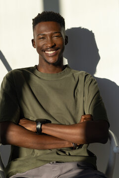African man sitting arms crossed smiling in olive T-shirt at plain wall sunlight showing smartwatch