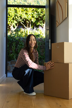 Asian woman crouching, touching stacked moving boxes in foyer by open front door with sunlit garden