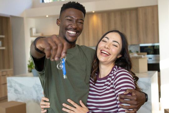 Diverse couple holding blue key fob, laughing and leaning at marble kitchen island unpacking boxes