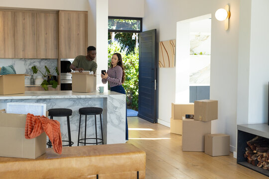 Diverse couple standing behind marble kitchen island unpacking moving boxes and holding smartphone