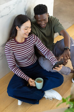 Diverse couple sitting on hardwood floor against marble wall, holding smartphone and mug