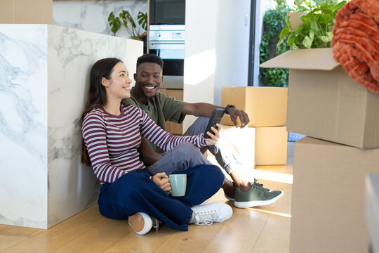 Couple sitting on hardwood floor next to marble island, holding phone, ceramic mug and moving boxes