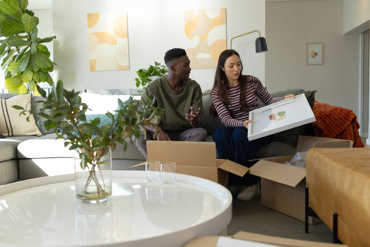 Diverse couple sitting at home holding framed artwork and discussing placement amid moving boxes