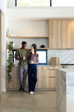 Diverse couple standing close together holding ceramic mugs while unpacking box in modern kitchen