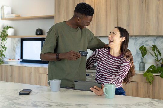 Diverse couple laughing while browsing online at kitchen island, holding tablet and credit card