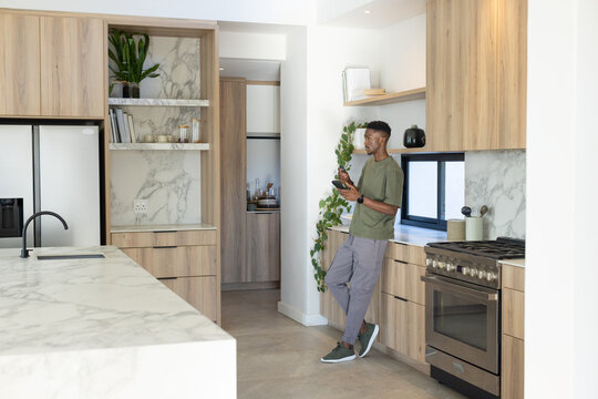 African American man leaning on counter in kitchen, holding smartphone by marble island, copy space