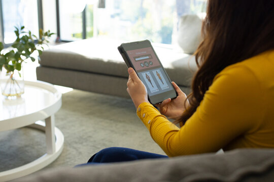 Adult woman sitting on gray sofa in yellow top holding tablet browsing styles home, copy space