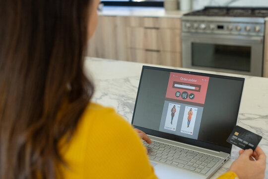 Woman in yellow sweater using laptop shopping at kitchen island, holding credit card, copy space